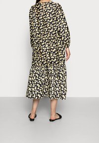 Schwarzes Kleid mit gelbem Blumenmuster, langen Ärmeln, geraffter Taille und gestuftem Rock. Aus leichtem Stoff gefertigt. Flache schwarze Sandalen getragen.