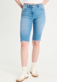 Bermudas en denim bleu avec une coupe ajustée, ourlets roulés, poches avant et zip. Associées à des baskets blanches et des chaussettes.