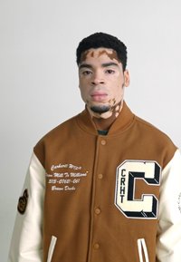 Smeđa jakna s varšavskim rukavima i kremastim rukavima, sa izvezenim logom "C" na prsima. Sadrži logo i tekstualne detalje na prednjoj strani.