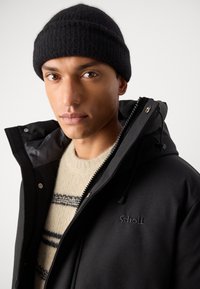 Black waterproof jacket with a zip front, adjustable hood, and logo detail; worn over a light textured sweater with dark stripes. Black knit beanie.