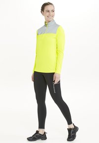 Langærmet, halv-zip aktiv top i neongul med grå skulderpaneler, parret med sorte leggings med et hvidt logo og reflekterende detaljer.