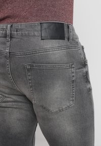 Vaqueros de mezclilla gris con un acabado ligeramente desgastado. Las características incluyen un parche de cuero negro en la cintura, dos bolsillos traseros y un diseño ajustado.