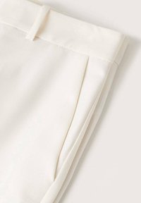 Pantalon sur mesure de couleur blanc cassé plié, montrant la ceinture, les passants de ceinture, la poche latérale et le détail du pli avant sur un tissu lisse.