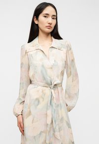 Μακρύ, αέρινο φόρεμα από διαφανές ύφασμα με απαλά παστέλ λουλουδένια σχέδια. Διαθέτει γιακά, μακριά μανίκια και λεπτομέρεια δεμένη στη μέση.