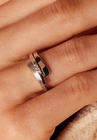 Twee in elkaar vergrendelde ringen om een vinger; de ene is zilver met een gladde afwerking, de andere is goud met een matte textuur en een rijtje kleine heldere stenen.