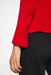 Red tailored jacket with a textured fabric and three red buttons on the cuff. Black pants with a pocket featuring a subtle heart-shaped stitch.