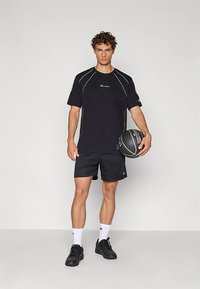 Černé atletické tričko s bílým kontrastním šitím, doplněné černými šortkami. Model drží basketbalový míč a má na sobě černé boty a bílé ponožky.