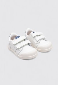 Zapatillas blancas con dos tiras de Velcro, textura de cuero suave, puntera redondeada y una etiqueta azul en la lengüeta. Suela de goma de color claro.