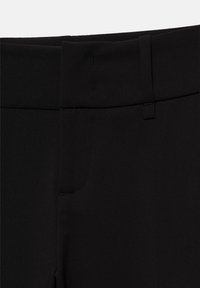 Gros plan sur la ceinture d’un pantalon noir avec des passants de ceinture et des boutonnières, montrant les détails des coutures et la texture du tissu.