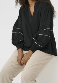 Blouse noire avec un col en V, des manches bouffantes ornées d'accents blancs en forme d'ondulations et une finition texturée. Portée avec un pantalon couleur crème.