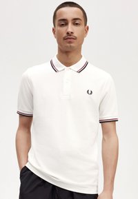 Witte polo met geribbelde kraag en manchetten met rode en marineblauwe strepen. Klein geborduurd logo op de borst. Zachte stof textuur.