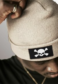 Tan ribbed beanie featuring a black patch with a white skull and crossbones design. The texture is knitted and close-fitted.