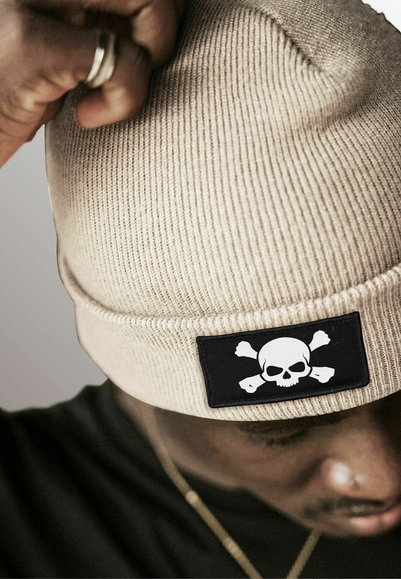 Tan ribbed beanie featuring a black patch with a white skull and crossbones design. The texture is knitted and close-fitted.