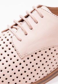 Chaussure en cuir rose clair avec des perforations carrées, des lacets assortis et des coutures visibles près du dessus et du panneau latéral.