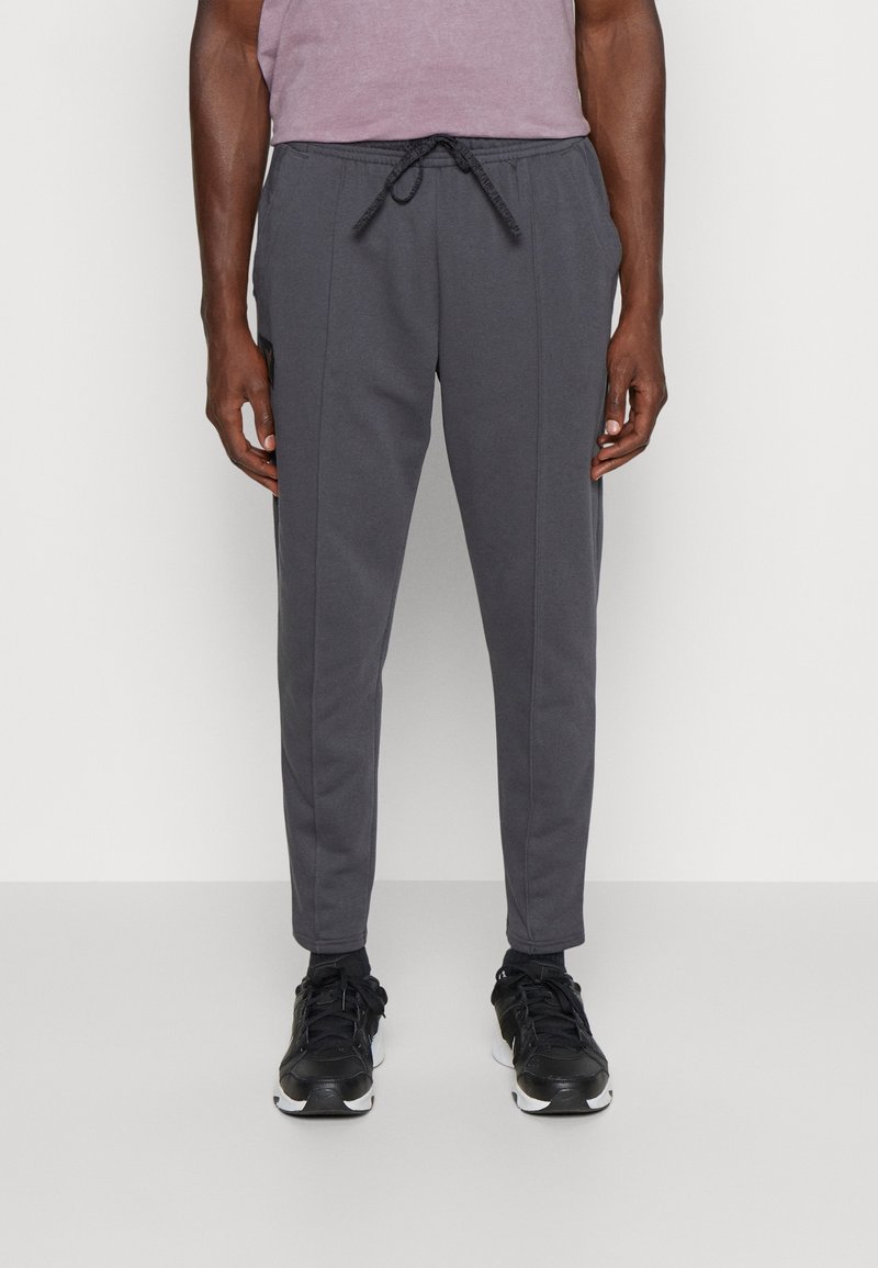 Hombre vistiendo pantalones jogger gris oscuro de corte ajustado con cintura elástica y cordón, combinados con zapatos deportivos negros y una camisa malva.