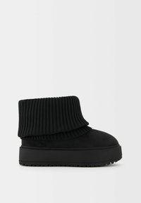 Bottine noire à la cheville avec un bord en tricot côtelé épais et une semelle plateforme épaisse, vue de côté sur un fond clair uni.
