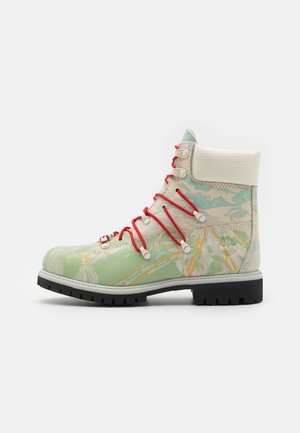 Hoge laars met groen-beige camouflagepatroon, rode veters, gewatteerde witte kraag, zwarte profielzool en Timberland-logo op de hiel.