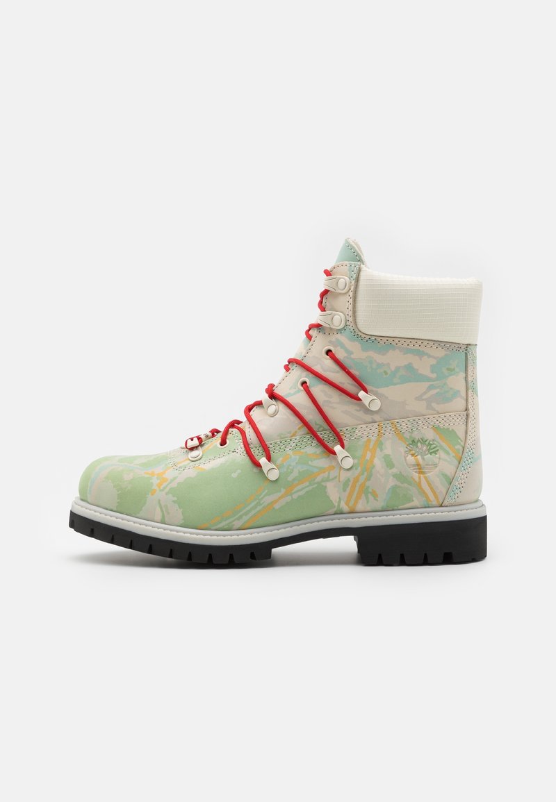 Botte haute avec motif camouflage vert et beige, lacets rouges, col blanc rembourré, semelle crantée noire et logo Timberland sur le talon.