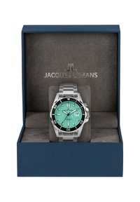 Montre en métal avec un cadran turquoise, un bracelet en acier inoxydable argenté et une lunette noire. Présentée dans une boîte bleue avec un intérieur en velours gris.