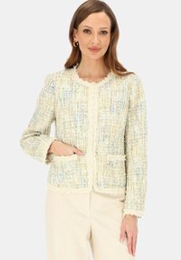 Kobieta ubrana w kremowo-niebieską tweedową kurtkę o teksturowanej strukturze z przednimi kieszeniami, w połączeniu z jasnobeżowymi spodniami, stojąca na jednolitym tle.