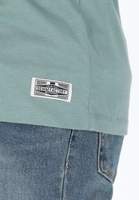 Ljusgrön bomullströja med en texturerad yta, med en vit etikett som står "ORIGINAL DENIM FACTORY" sydd på fållen.