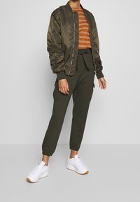 Olivgrön bomberjacka över en topp med orange ränder, parat med olivgröna cargobyxor och vita sneakers med gummisulor.