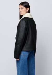 Giacca nera con finitura testurizzata e colletto in pelliccia sintetica beige. Presenta un design morbido e viene abbinata a jeans blu a vita alta.