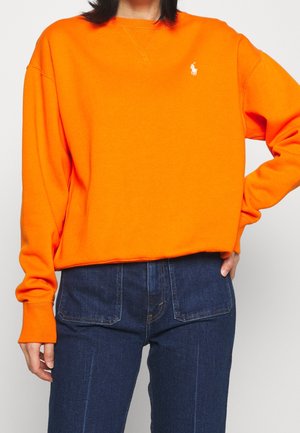 Person wearing a bright orange sweatshirt with a small white logo on the chest and dark blue denim jeans, hand on hip.