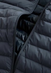 Geacă puffer neagră cu textură matlasată, cu un fermoar neted și căptușeală interioară. Materialul pare ușor și izolat.