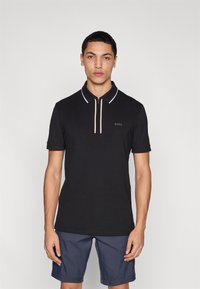 Polo noir avec col zippé, accents blancs et beiges, manches courtes. Présente un logo discret sur la poitrine. Associé à un short bleu.