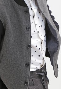 Grijs wollen blend vest met zwarte knopen over een witte blouse met zwarte stippen. De outfit omvat donker op maat gemaakte broek met een koord.