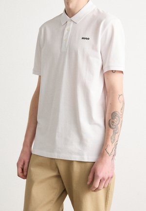Personne portant un polo blanc Hugo et un pantalon beige, avec un tatouage de serpent visible sur l'avant-bras droit.