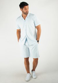 Chemise bleu clair à manches courtes avec boutons et manches retroussées, associée à un short assorti. Texture lisse, design simple, et chaussures blanches.