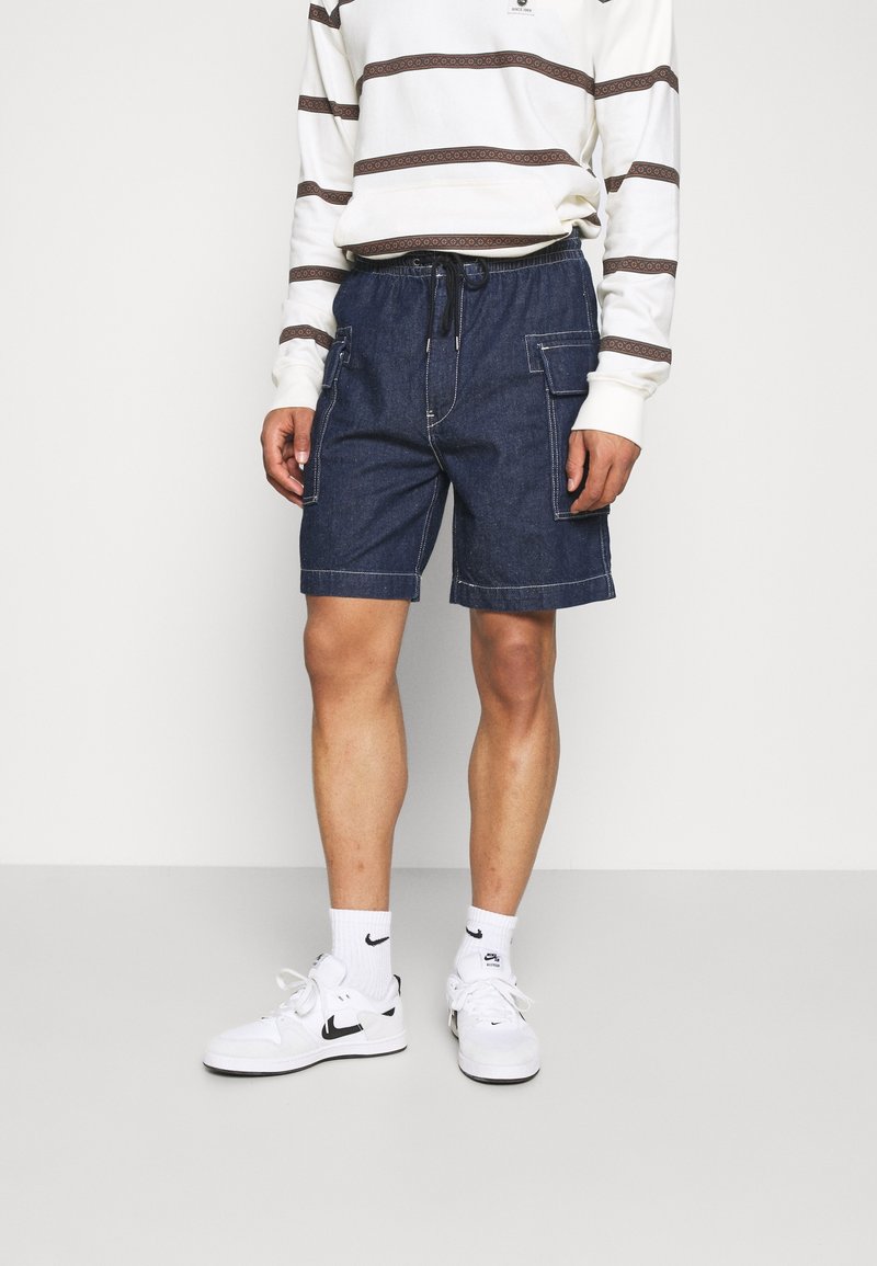 Shorts cargo en denim bleu foncé avec surpiqûres blanches contrastantes, dotés de multiples poches latérales et d'une taille élastique avec cordon de serrage.
