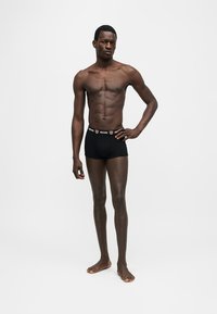 Λεπτός άνδρας στέκεται ξυπόλυτος, φορώντας μαύρα boxer Moschino με λογότυπο και εικονίδια αρκουδάκι στη μέση, πάνω σε λευκό φόντο.