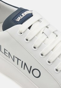 Fehér bőr sneaker tengerészkék kiegészítőkkel, texturált nyelvvel, fehér fűzőkkel és fekete "VALENTINO" logóval az oldalán.