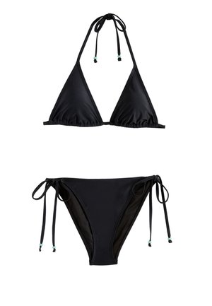 Frau im schwarzen Bikini läuft barfuß am Sandstrand nahe dem Ozean und hält eine grüne Kokosnuss mit einem Strohhalm.