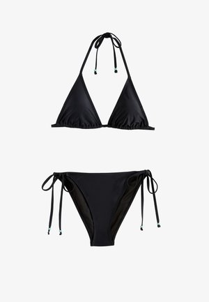Frau im schwarzen Bikini läuft barfuß am Sandstrand nahe dem Ozean und hält eine grüne Kokosnuss mit einem Strohhalm.