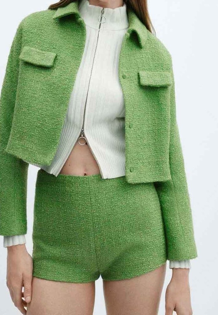Femme portant une veste courte texturée verte et un short taille haute assorti par-dessus un pull court blanc zippé.