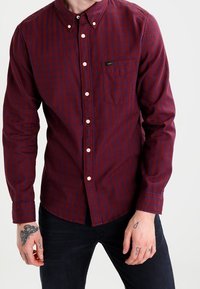 Camicia a quadri maroon e blu navy con maniche lunghe, colletto, tasca frontale e bottoni bianchi, realizzata in tessuto morbido.