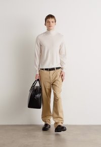 Pull en col roulé crème, pantalon beige, ceinture noire et sac en cuir. Tissu lisse, coupe décontractée, et chaussures noires épaisses. Fond uni.