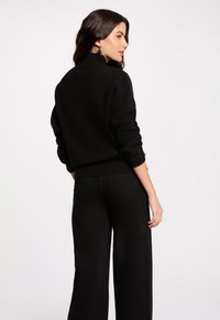 Jersey negro de punto con cuello alto, que presenta textura acanalada y mangas enrolladas, combinado con pantalones negros de talle alto y pernera ancha.