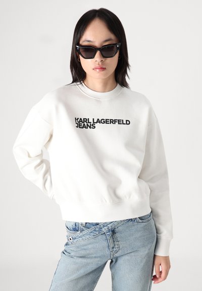 Бял суитшърт с черен текст "KARL LAGERFELD JEANS", с рибени маншети и подгъв, съчетан с светлосини дънки. Моделът носи черни слънчеви очила.