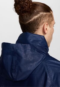 Chaqueta azul marino con un cuello alto, que presenta un patrón texturizado. El material parece suave, ligero y resistente al agua.