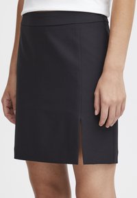 Rovná černá sukně ke kolenům se bočním rozparkem, kterou nosí osoba s uvolněnýma rukama a světlým tónem pleti.