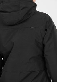 Svart utomhusjacka med texturerad tyg, huva och en mittsöm. Har en liten logotagg på den övre delen av ryggen.