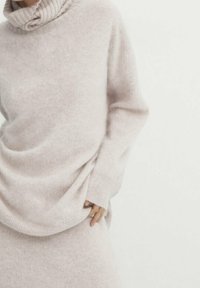 Ljus beige stickad tröja med ribbade krage och lös passform. Mjuk textur, långa ärmar och subtilt draperingsdetalj vid midjan.