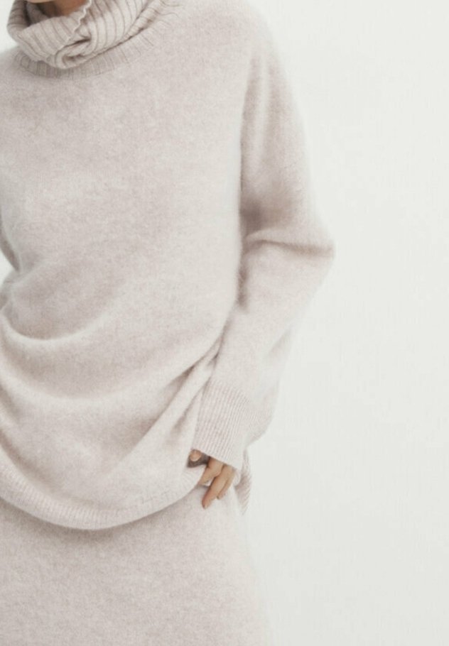 Ljus beige stickad tröja med ribbade krage och lös passform. Mjuk textur, långa ärmar och subtilt draperingsdetalj vid midjan.