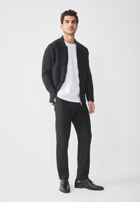 Chaqueta negra con cremallera sobre una camiseta blanca, combinada con pantalones negros y zapatos negros. Texturas suaves, diseño minimalista y un ajuste relajado.