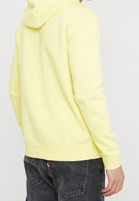 Žlutá mikina s kapucí a měkkou texturou, s klokankou a žebrovanými manžetami, kombinovaná s tmavě šedými džíny.